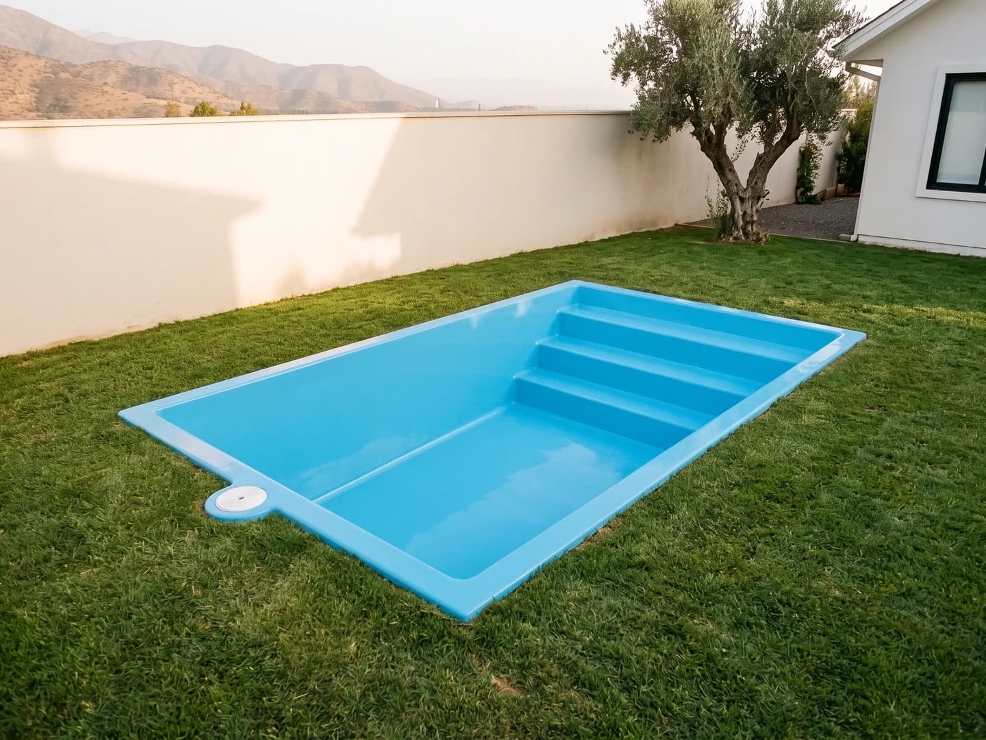 Piscina de fibra de vidrio de 3,50 por 2,50 metros instalada a nivel del pasto en el jardín de una casa, vista desde arriba, vacía y sin borde, con la aleta de fibra a la vista y la escalera de tres peldaños a lo ancho de un extremo