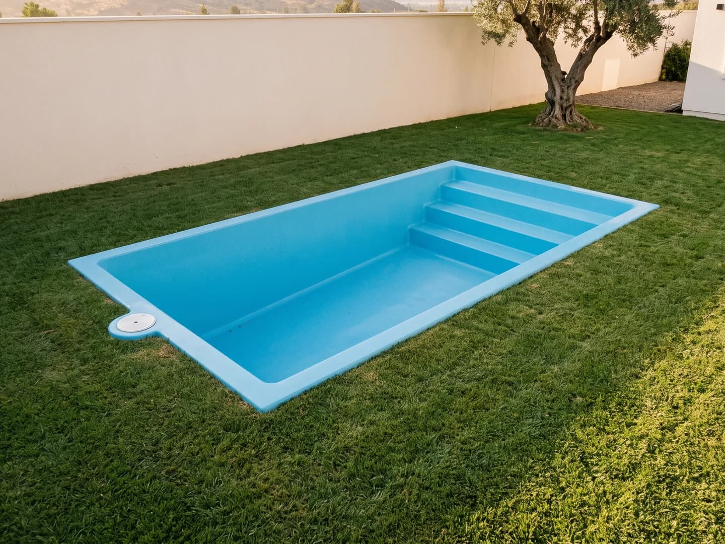 Piscina de fibra de vidrio de 4,00 por 2,50 metros instalada a nivel del pasto en el jardín de una casa, vista desde arriba, vacía y sin borde, con la aleta de fibra a la vista y la escalera de tres peldaños a lo ancho de un extremo