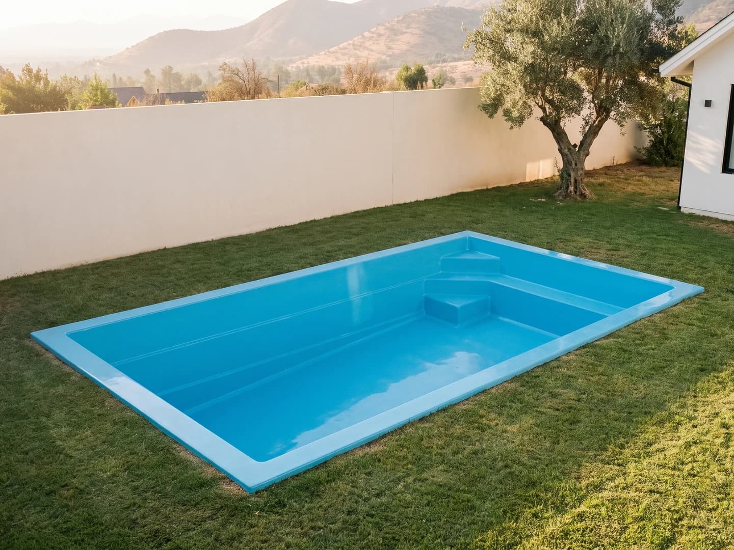 Piscina de fibra de vidrio de 6,00 por 3,00 metros instalada a nivel del pasto en el jardín de una casa, vista desde arriba, vacía y sin borde, con la aleta de fibra a la vista y un bloque de tres peldaños en una esquina del extremo bajo