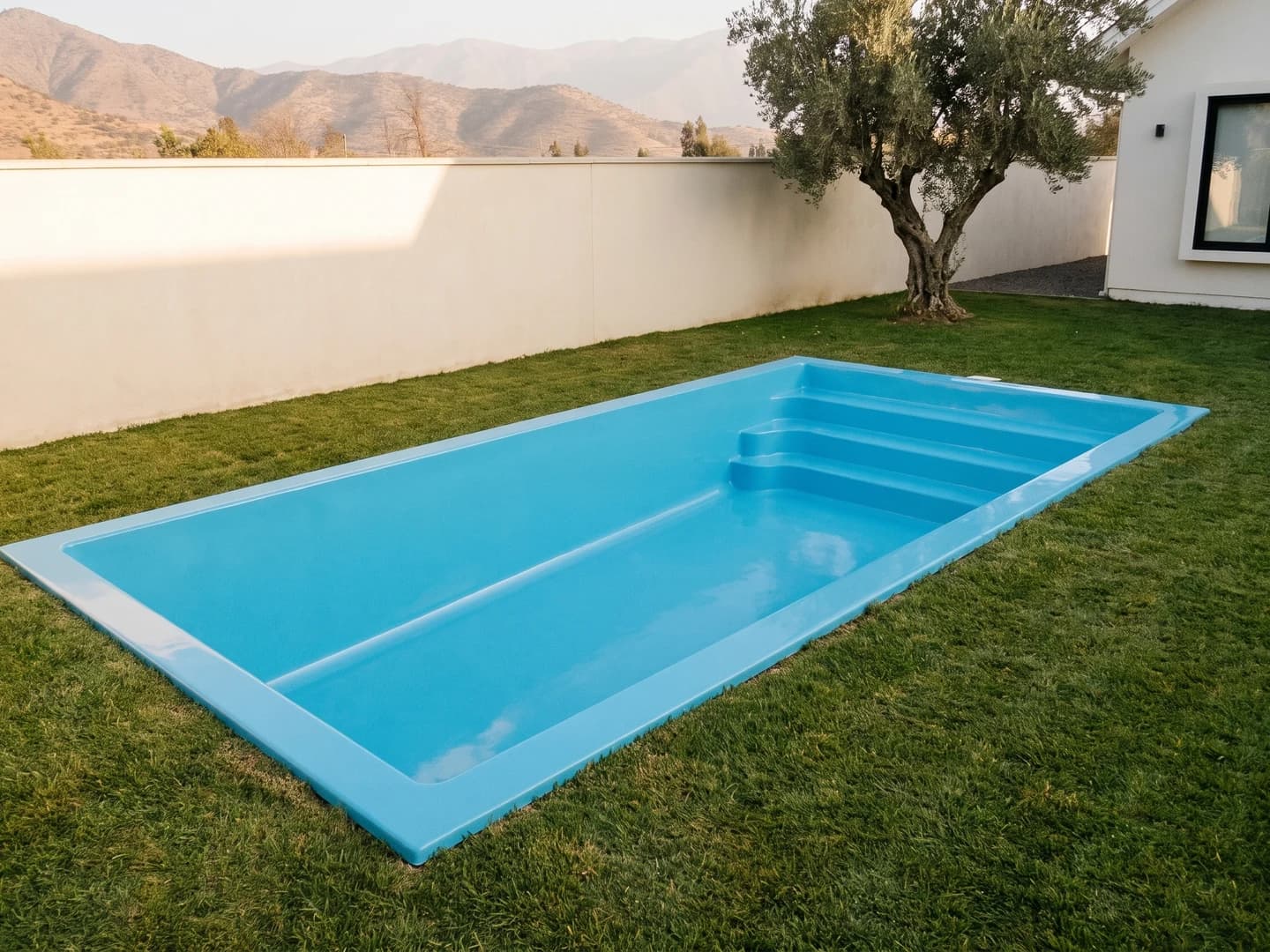 Piscina de fibra de vidrio de 7,00 por 3,10 metros instalada a nivel del pasto en el jardín de una casa, vista desde arriba, vacía y sin borde, con la aleta de fibra a la vista y una escalera de tres peldaños de frente curvo a lo ancho de un extremo