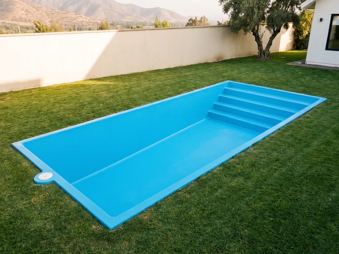 Piscina de fibra de vidrio de 9,00 por 4,00 metros instalada a nivel del pasto en el jardín de una casa, vista desde arriba, vacía y sin borde, con la aleta de fibra a la vista, la tapa redonda del skimmer en un costado y la escalera de cuatro peldaños a lo ancho de un extremo